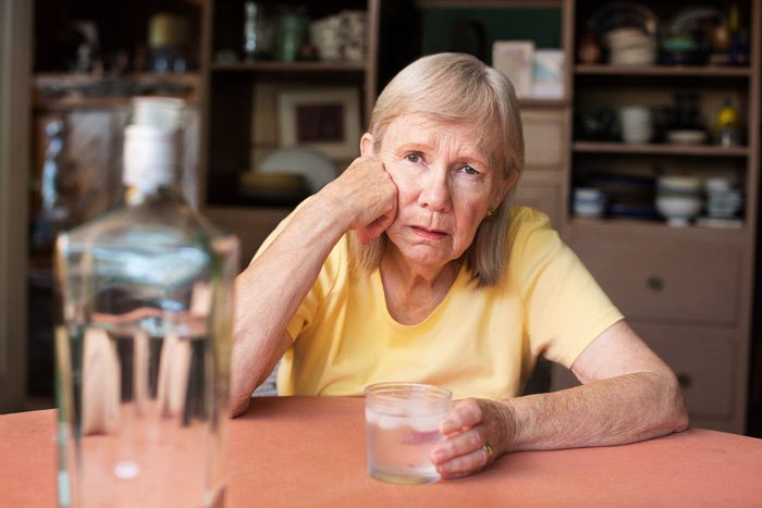 senior woman sitting at a table at home drinking liquor and looking sad - loneliness