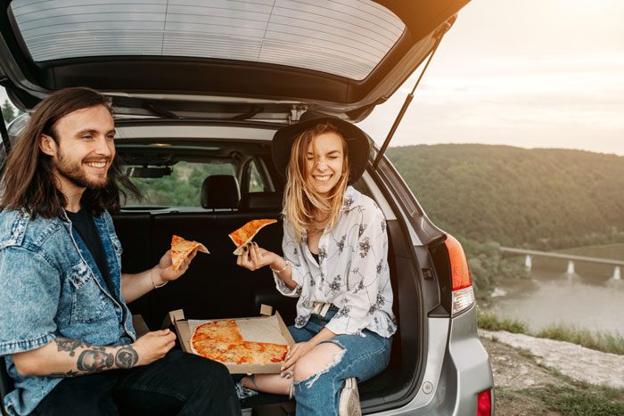 attractive and hip young couple eating sliced pizza out of the back of a hatchback on a road trip - sober vacation